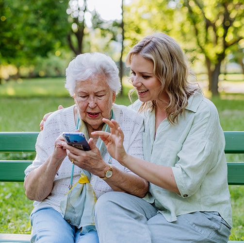Granddaughter-Helping-Grandma-With-Phone-On-Park-Bench.jpg